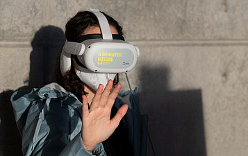 Persona joven de frente y medio cuerpo, con gafas de realidad virtual y la mano derecha hacia delante como tocando algo. Produce intriga.