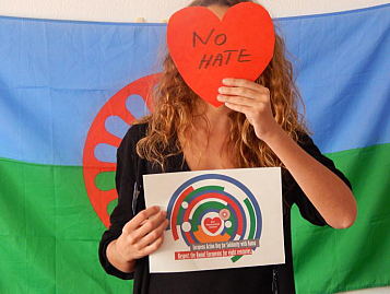 mujer con bandera gitana al fondo y un corazón de papel en la cara con "no hate"