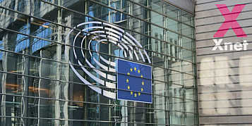 Fotografia de cristalera del parlament europeu amb la bandera i a la dreta el logo de X-net