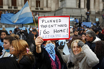 Mujeres manifestándose con una pancarta que dice: "Planned parent hood ¡Go Home! Lezica 3902 Almagro" Se ven banderas argentinas entre la multitud. En primer plano las mujeres que la sostienen están contentas y activas.