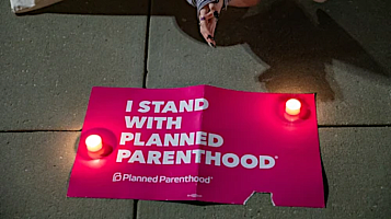 pink poster on the street floor with two candles and in the top of the image, the hands of a woman. the poster: pink and it says: i stand with planned parenthood